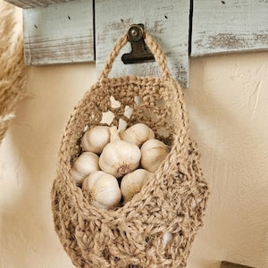 May include: A jute basket filled with garlic bulbs hangs from a rustic wooden shelf. The basket is suspended by a jute handle and secured with a metal clip. The garlic bulbs are white with brown roots, and the basket has an open weave design.