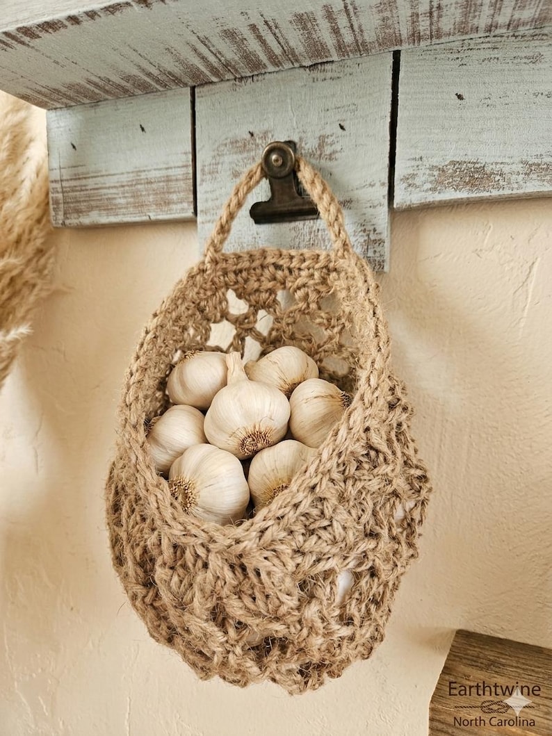 May include: A jute basket filled with garlic bulbs hangs from a rustic wooden shelf. The basket is suspended by a jute handle and secured with a metal clip. The garlic bulbs are white with brown roots, and the basket has an open weave design.