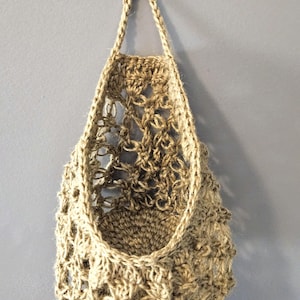 May include: A hanging woven basket made from natural jute fibre. The basket has an open, airy design with a loop at the top for hanging. The basket is empty and hangs against a grey wall.