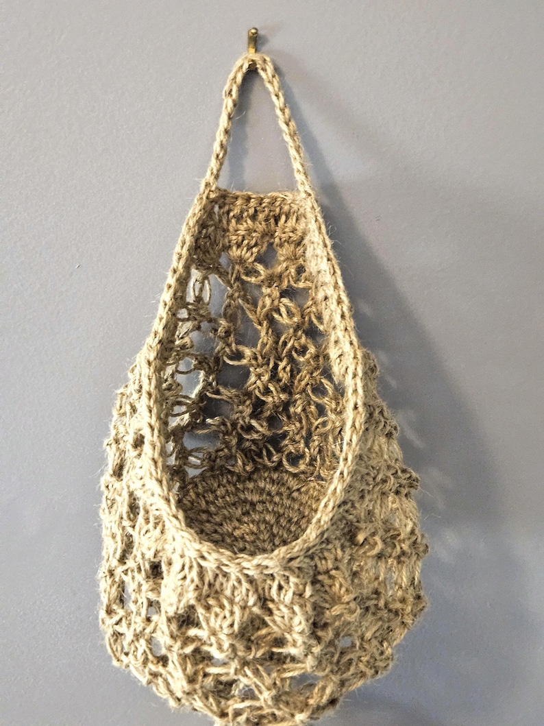 May include: A hanging woven basket made from natural jute fibre. The basket has an open, airy design with a loop at the top for hanging. The basket is empty and hangs against a grey wall.