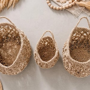 May include: Three woven baskets of different sizes, each with a handle. The baskets are made from a light brown, natural fibre and have an open weave design. The baskets are empty and are hanging against a neutral background.