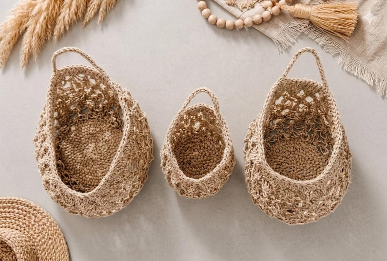 May include: Three woven baskets of different sizes, each with a handle. The baskets are made from a light brown, natural fibre and have an open weave design. The baskets are empty and are hanging against a neutral background.
