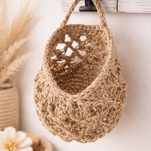 May include: A hanging jute basket, woven with a natural tan-coloured fibre. The basket is suspended from a black metal clip. The open weave design is suitable for storage and adds a rustic touch to any room.