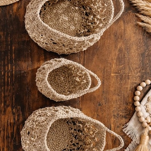 May include: Three handmade crochet baskets in varying sizes, arranged on a wooden surface. The baskets are a natural beige colour, with a textured, open weave design. The handles are integrated into the basket's structure. Decorative elements include dried grasses and wooden beads.