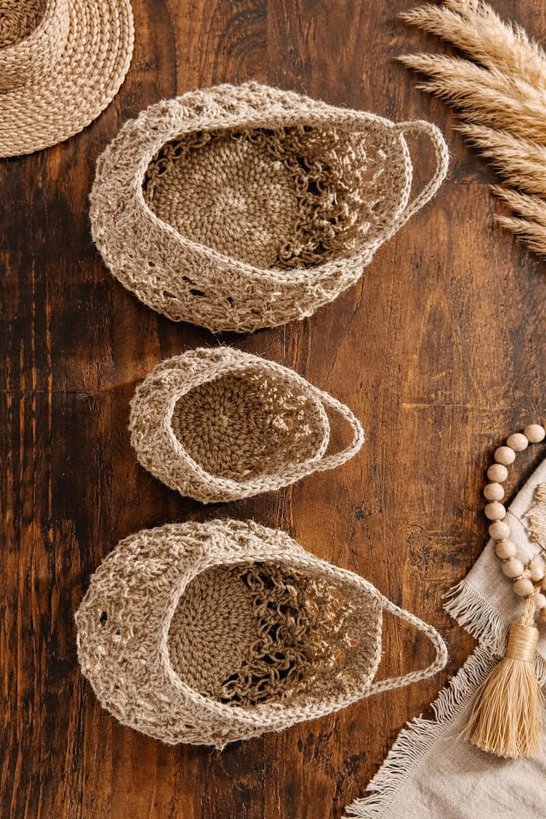 May include: Three handmade crochet baskets in varying sizes, arranged on a wooden surface. The baskets are a natural beige colour, with a textured, open weave design. The handles are integrated into the basket's structure. Decorative elements include dried grasses and wooden beads.