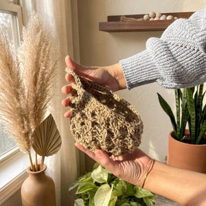 May include: A hand-crocheted, beige basket with an open weave design. The basket is held in a person's hands, highlighting its shape and texture. The background features a vase with dried plants and other decorative items.