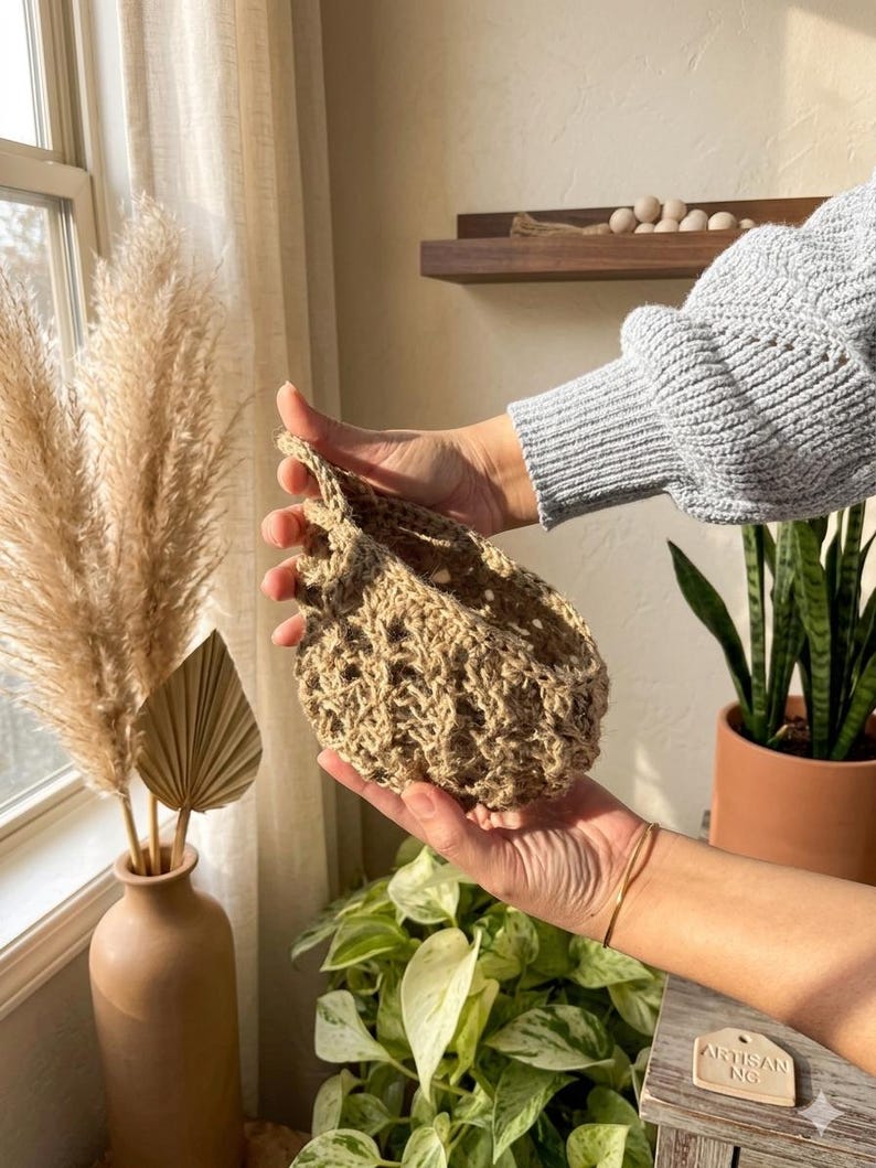May include: A hand-crocheted, beige basket with an open weave design. The basket is held in a person's hands, highlighting its shape and texture. The background features a vase with dried plants and other decorative items.