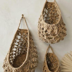 May include: Three jute hanging baskets in different sizes are shown against a neutral wall. The baskets have a natural, earthy colour and an open weave design. Each basket is suspended by a loop, offering a rustic aesthetic.