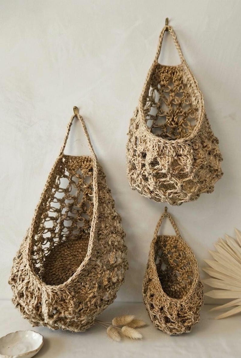 May include: Three jute hanging baskets in different sizes are shown against a neutral wall. The baskets have a natural, earthy colour and an open weave design. Each basket is suspended by a loop, offering a rustic aesthetic.