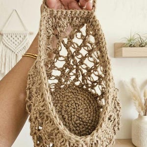 May include: A woven jute basket with a handle, held in a hand. The basket features an open, net-like weave and a solid, circular base. The natural brown jute gives the basket a rustic appearance. The basket is designed for storage or display, with a handle for hanging.
