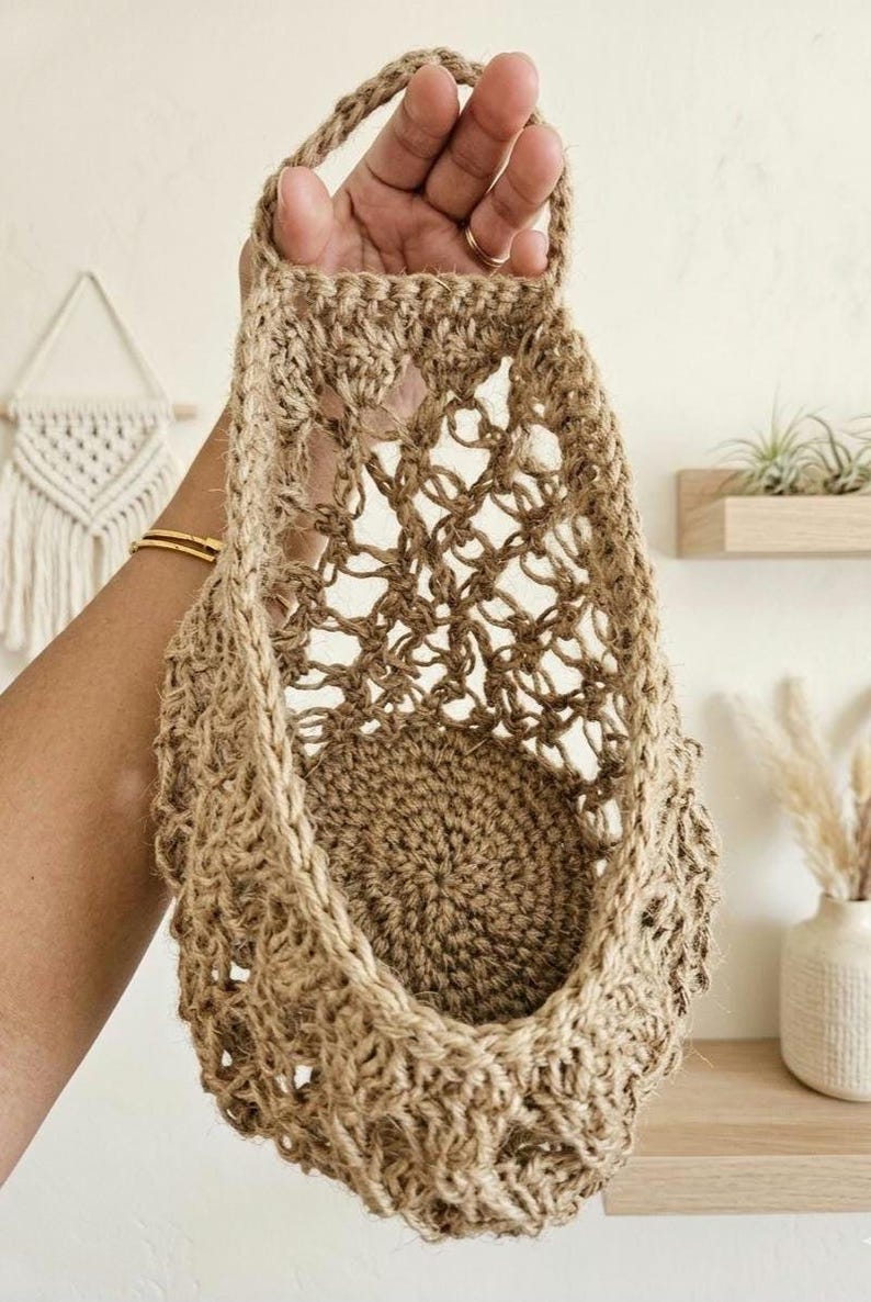 May include: A woven jute basket with a handle, held in a hand. The basket features an open, net-like weave and a solid, circular base. The natural brown jute gives the basket a rustic appearance. The basket is designed for storage or display, with a handle for hanging.