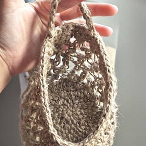 May include: A small, open-weave basket made of natural-coloured jute fibre. The basket has a round base and a handle made of the same material. The basket is held by a hand, and the background is blurred.