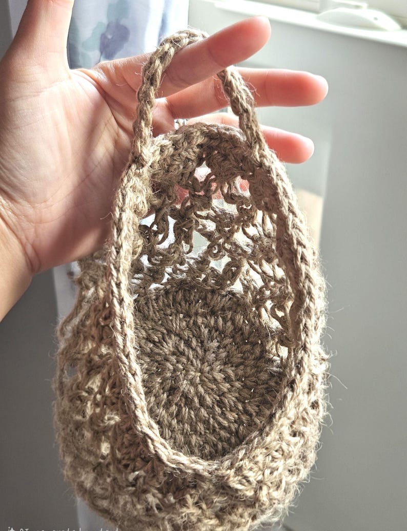 May include: A small, open-weave basket made of natural-coloured jute fibre. The basket has a round base and a handle made of the same material. The basket is held by a hand, and the background is blurred.