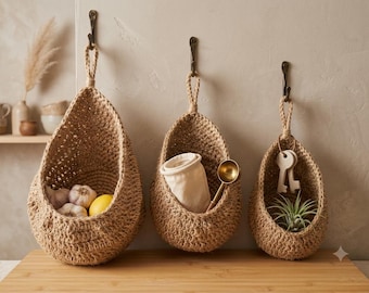May include: Three teardrop-shaped hanging baskets made of woven tan jute fiber. Each basket features a thick braided loop for suspension from a metal hook. The baskets contain garlic bulbs, a lemon, a fabric pouch, a gold spoon, and a small plant.
