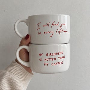 Puede incluir: Dos tazas de cerámica blanca apiladas verticalmente. La taza superior tiene el texto rojo "I will find you in every lifetime". La taza inferior tiene el texto rojo "MY GIRLFRIEND IS HOTTER THAN MY COFFEE". Ambas tazas tienen un asa redonda.