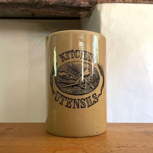May include: A cylindrical beige ceramic utensil holder with a glossy finish. The front features a black printed graphic of crossed kitchen tools inside an oval, surrounded by the text KITCHEN UTENSILS in a bold, serif font with wheat stalks.