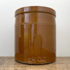 May include: A cylindrical brown glazed stoneware crock featuring a raised beaded band detail around the upper section and the embossed text 48 OZ near the base. The container has a smooth, glossy finish and a flat rim.