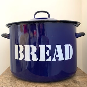 May include: A dark blue enamel bread bin with a black rim and handles. The word "BREAD" is stencilled in white across the front. The bin has a lid with a black handle. It is on a wooden surface.