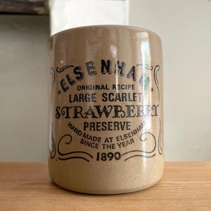 May include: A cylindrical beige stoneware crock featuring black printed text that reads ELSENHAM ORIGINAL RECIPE LARGE SCARLET STRAWBERRY PRESERVE HAND MADE AT ELSENHAM SINCE THE YEAR 1890, framed by decorative black scrollwork lines on the front surface.