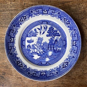 May include: A blue and white ceramic dinner plate featuring a willow pattern design. The plate depicts a traditional Chinese landscape with a bridge, trees, and a pagoda.
