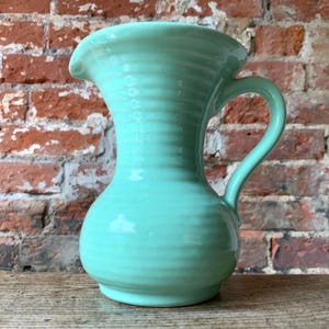 May include: A pale green ceramic jug with a handle and a spout. The jug has a ribbed texture and a rounded base. The background is a brick wall and a wooden surface.
