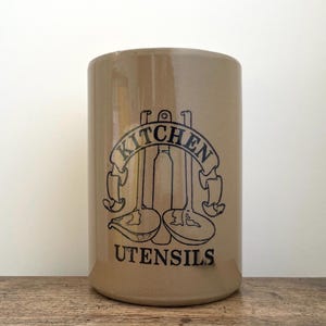May include: Tan ceramic kitchen utensil holder with a black graphic of kitchen utensils and the words "KITCHEN UTENSILS". The cylindrical container is on a wooden surface.