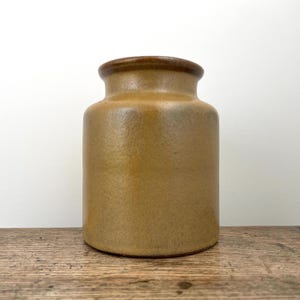 May include: A cylindrical stoneware jar with a textured, matte mustard-yellow finish and a smooth, dark brown rim. The vessel features a wide, short neck and a flat base, showcasing a consistent, earthy tone throughout its simple, functional form.
