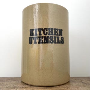 May include: A cylindrical beige stoneware crock with a smooth, speckled finish. The front features the words KITCHEN UTENSILS printed in bold, black, distressed-style capital letters. The container has a flat base and an open top design.