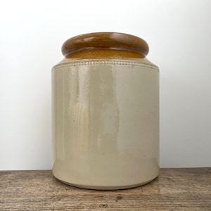 May include: A cylindrical stoneware crock featuring a smooth, cream-coloured body and a contrasting glossy brown glazed rim. The upper section of the cream-coloured body is decorated with a subtle, embossed beaded texture encircling the vessel.