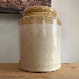 May include: A cylindrical ceramic crock featuring a glossy, speckled cream-coloured body and a distinct, smooth tan-coloured rim. The upper section of the vessel includes a subtle, raised beaded texture detail encircling the neck area.