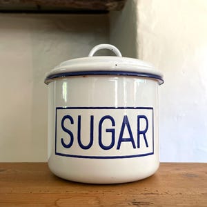 May include: A white enamel canister with a matching lid and a blue rim. The front features a rectangular blue border enclosing the word SUGAR in bold blue capital letters. The container has a simple arched handle on top of the lid.