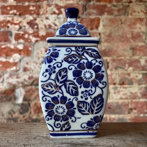May include: A white porcelain jar with a blue and gold floral design. The jar has a lid and is decorated with a repeating pattern of flowers and leaves.