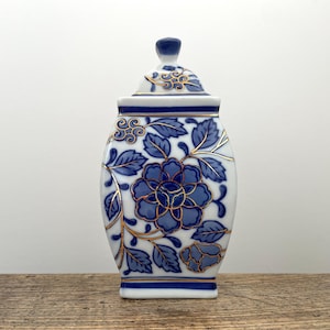 May include: A white ceramic jar with a lid, featuring a blue floral pattern and gold-coloured outlines. The jar has a rectangular body with rounded sides, decorated with blue leaves and flowers, and a matching lid with a small knob on top.