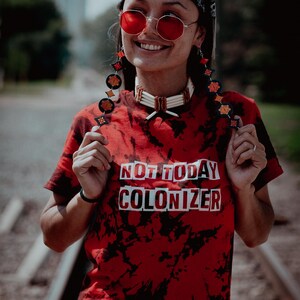 Könnte beinhalten: Eine Person trägt ein rotes Batik-Shirt mit dem aufgedruckten Text "NOT TODAY COLONIZER" in Weiß. Sie trägt ein schwarzes Bandana, eine rote Sonnenbrille und Perlenohrringe.