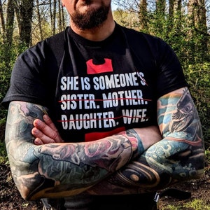 Peut inclure: Une personne portant un t-shirt noir avec le texte "She is someone's sister. mother. daughter. wife." avec des lignes tirées sur les mots "mother", "daughter" et "wife".