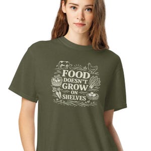 Puede incluir: Camiseta verde oliva con el texto blanco "FOOD DOESN'T GROW ON SHELVES". El diseño incluye ilustraciones de animales de granja, verduras y un nido. Una camiseta cómoda, perfecta para quienes se interesan por los alimentos sostenibles.