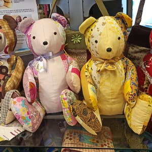 May include: Four patchwork teddy bears, each made from different fabrics and colours, are displayed on a glass shelf. The bears are sitting upright, with their arms outstretched. The bears are all wearing a ribbon around their necks. The bears are all handmade and unique.