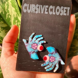 May include: Two blue hands with pink and white eyes and red pupils. The hands are holding a black card with the text "CURSIVE CLOSET" in white.