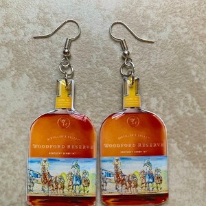 May include: A pair of dangle earrings featuring a Woodford Reserve bottle design. The earrings have a silver hook and a clear, rectangular top. The bottle design includes the words "Woodford Reserve" and a Kentucky Derby scene.