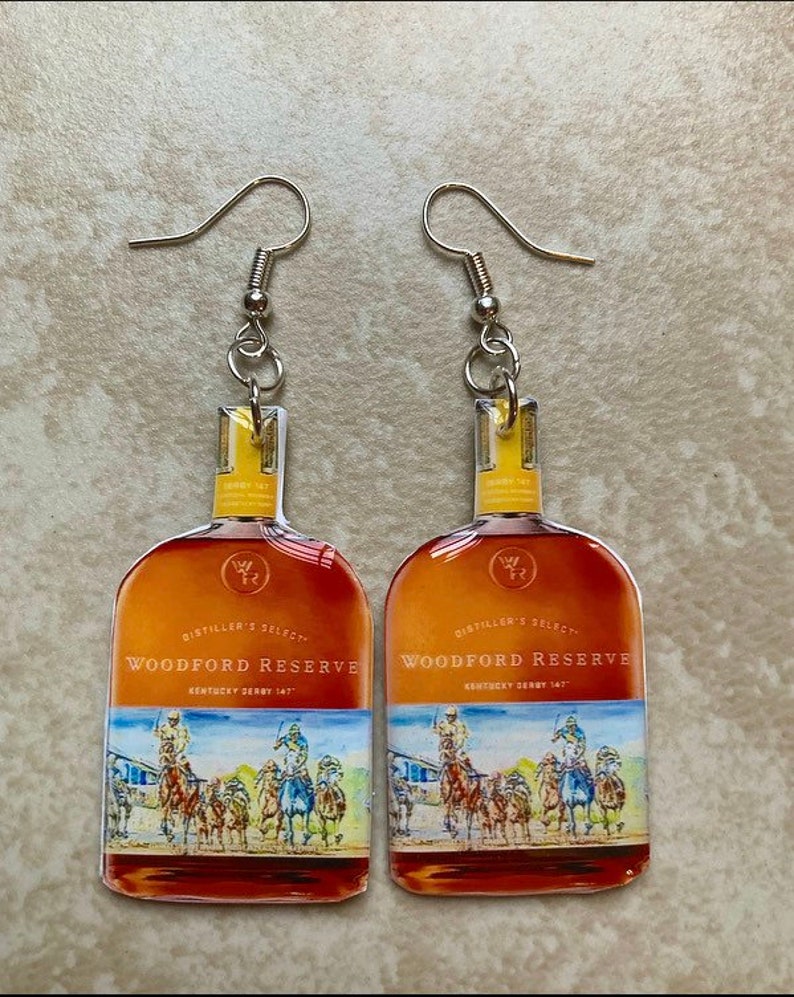 May include: A pair of dangle earrings featuring a Woodford Reserve bottle design. The earrings have a silver hook and a clear, rectangular top. The bottle design includes the words "Woodford Reserve" and a Kentucky Derby scene.
