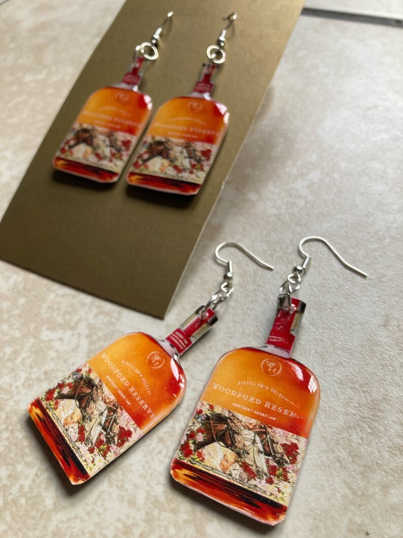 May include: Earrings featuring a Woodford Reserve bottle design. The earrings have a silver hook and a rectangular shape with an orange and red gradient. The bottle design includes text and a horse racing scene with red roses.
