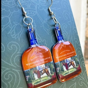 May include: A pair of earrings shaped like Woodford Reserve bourbon bottles. The earrings have a blue and brown color scheme, with a picture of a horse race on the front. The earrings are attached to silver-colored fish hook ear wires.