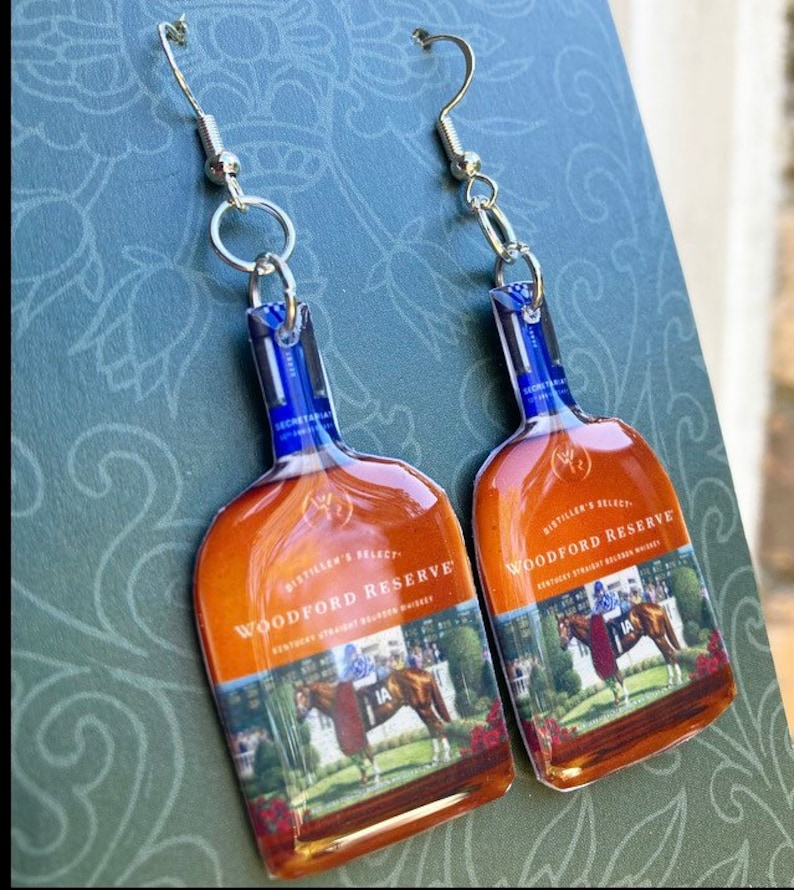 May include: A pair of earrings shaped like Woodford Reserve bourbon bottles. The earrings have a blue and brown color scheme, with a picture of a horse race on the front. The earrings are attached to silver-colored fish hook ear wires.