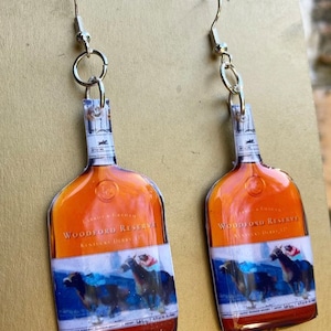 May include: A pair of dangle earrings featuring a Woodford Reserve bottle design. The earrings have a silver-colored hook and a clear plastic bottle shape with a label that includes the words "Woodford Reserve Kentucky Derby". The bottle design has a brown and blue color scheme.
