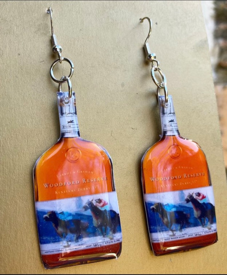 May include: A pair of dangle earrings featuring a Woodford Reserve bottle design. The earrings have a silver-colored hook and a clear plastic bottle shape with a label that includes the words "Woodford Reserve Kentucky Derby". The bottle design has a brown and blue color scheme.