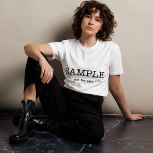 May include: A white t-shirt with black text that reads "SAMPLE not for sale". The person wearing the shirt is sitting on a marble floor.