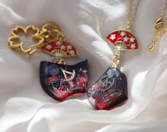 May include: Two gold-tone charms with red and white fan-shaped pendants and black resin charms with red and gold accents. The charms are attached to a gold chain and a gold key ring.