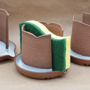 May include: A handmade, brown ceramic sponge holder with a scalloped top and a white-painted rim. A green and yellow sponge sits inside the holder. The holder is on a matching saucer. Two other holders are visible in the background.