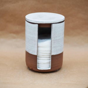 May include: A ceramic cotton pad holder with a cylindrical shape. The bottom section is a reddish-brown color, while the top and lid are white. A vertical slot reveals the cotton pads inside. The holder is designed for bathroom or vanity use.