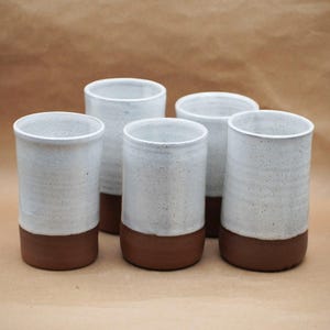 May include: A set of five cylindrical ceramic tumblers featuring a speckled white glaze on the upper portion and a solid, unglazed reddish-brown clay base. Each vessel has a smooth, straight-sided profile with a simple, functional design.